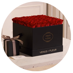 Black square box filled with red roses labeled 'VENUS ET FLEUR' next to a smaller black gift box tied with a brown ribbon, placed on a white surface.